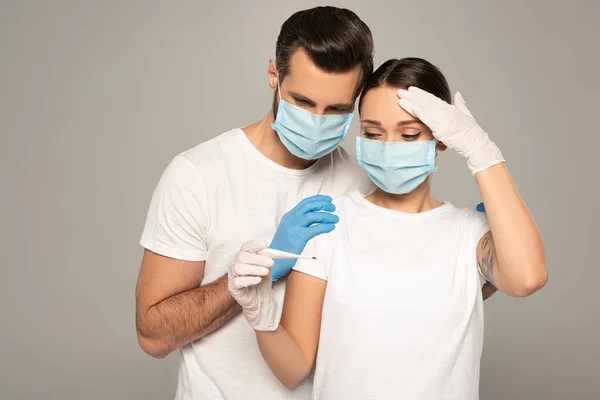 Man in latex gloves hugging girlfriend with thermometer and hand near forehead isolated on grey — Stock Photo