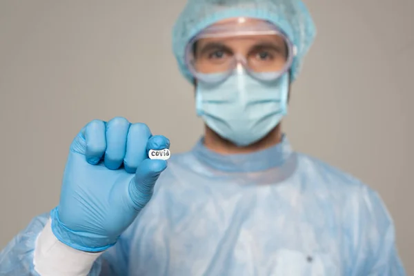 Enfoque selectivo del médico en googles de seguridad y píldora de retención de máscara médica con letras covid aisladas en gris - foto de stock