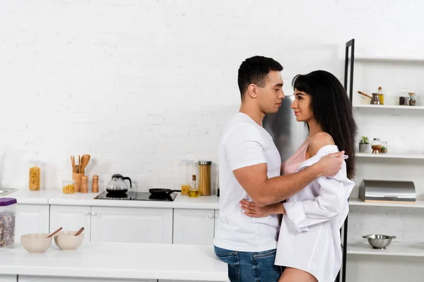 Pareja interracial mirándose, sonriendo y abrazándose cerca de la mesa en la cocina - foto de stock