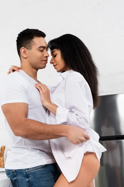 Mujer afroamericana con los ojos cerrados sonriendo y abrazando al hombre en la cocina - foto de stock