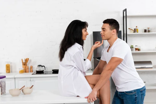 Afroamerikanerin sitzt auf Tisch, lächelt und schaut Freund in Küche an — Stockfoto