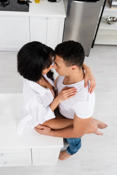 Vista de ángulo alto de la mujer afroamericana sentada en la mesa y abrazándose con su novio en la cocina - foto de stock