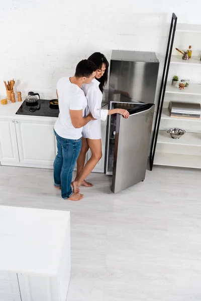 Vista de alto ângulo do homem abraçando mulher afro-americana perto de geladeira na cozinha — Fotografia de Stock