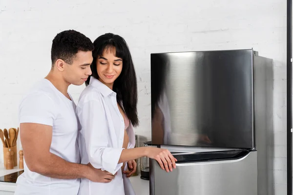 Homem abraçando mulher afro-americana perto de geladeira na cozinha — Fotografia de Stock