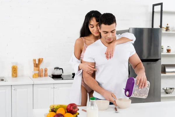 Homem com mulher afro-americana sorrindo e colocando cereal café da manhã em tigelas perto da mesa na cozinha — Fotografia de Stock