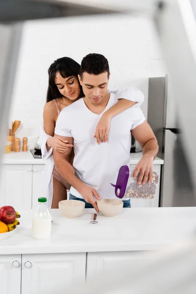 Selective focus of interracial couple hugging and man putting breakfast cereal in bowls near table in kitchen — Stock Photo