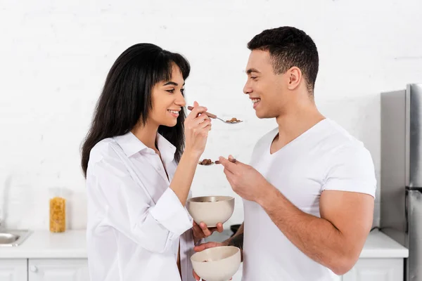 Casal interracial alimentando uns aos outros e sorrindo na cozinha — Fotografia de Stock