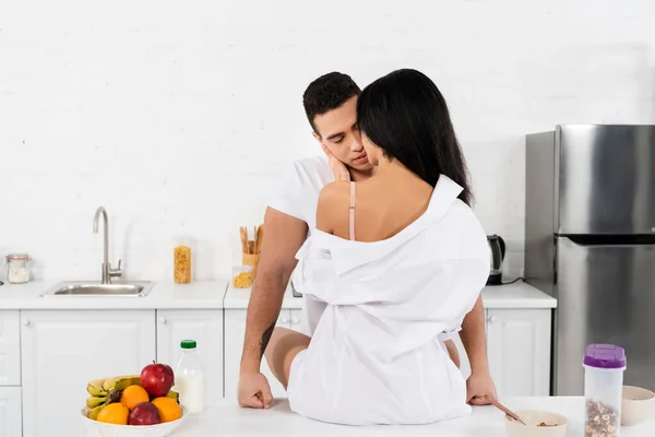 African american girl sitting on table, hugging and kissing man in kitchen — Stock Photo
