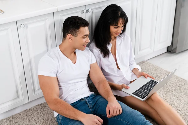 Vue grand angle du couple interracial souriant et regardant l'ordinateur portable près des armoires de cuisine sur le sol — Photo de stock