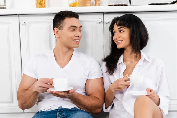 Couple interracial avec des soucoupes et des tasses de café souriant et se regardant les uns les autres près des armoires de cuisine — Photo de stock