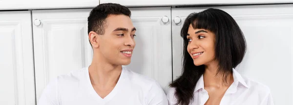 Interracial Paar lächelt und schaut einander in der Nähe von Küchenschränken, Panoramaaufnahme — Stockfoto