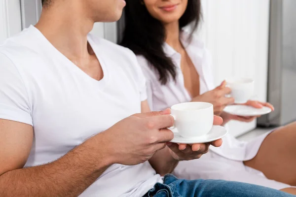 Vista recortada de pareja interracial con platillos y tazas de café en la cocina - foto de stock