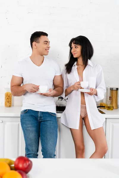 Foco seletivo de casal interracial com pires e xícaras de café sorrindo e olhando um para o outro na cozinha — Fotografia de Stock