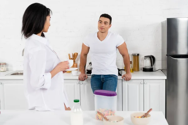 Hombre cerca de gabinetes de cocina sonriendo y mirando a la chica afroamericana con platillo y taza de café cerca de la mesa - foto de stock