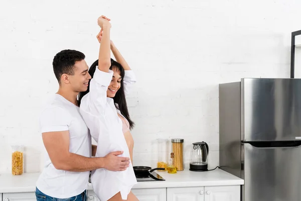 Uomo che abbraccia felice ragazza africana americana con le mani in aria da dietro in cucina — Foto stock
