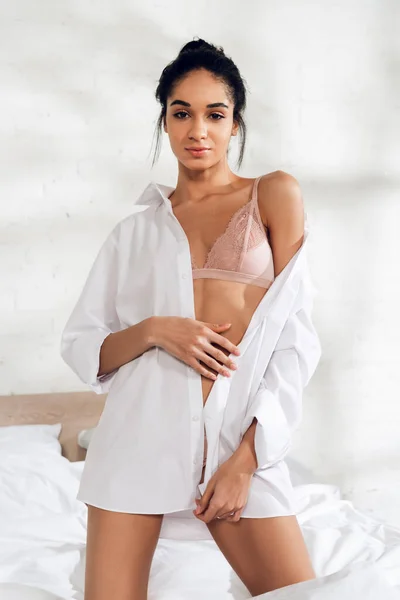 Sexy african american woman in white shirt looking at camera on bed in bedroom — Stock Photo