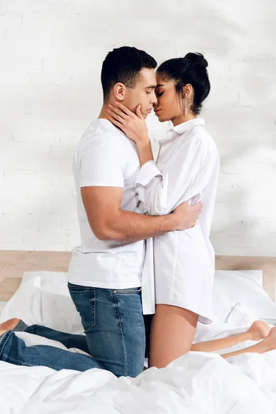 Interracial couple hugging and kissing on knees on bed in bedroom — Stock Photo