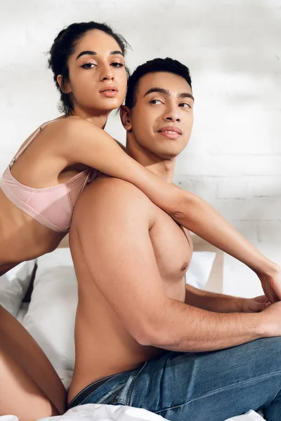 Afro-americana mujer abrazando novio por detrás y mirando la cámara en el dormitorio - foto de stock