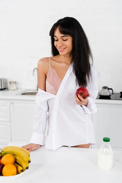 Mujer afroamericana sosteniendo manzana y sonriendo cerca de la mesa en la cocina - foto de stock