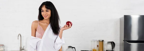 Ragazza afroamericana che tiene la mela, sorride e distoglie lo sguardo in cucina, colpo panoramico — Foto stock