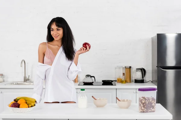 Afroamerikanerin hält Apfel in der Hand, lächelt und schaut am Tisch in der Küche weg — Stockfoto