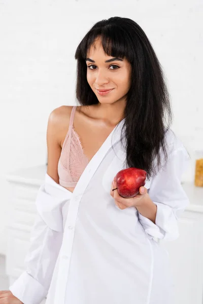 Africano chica americana con manzana sonriendo y mirando a la cámara en la cocina - foto de stock