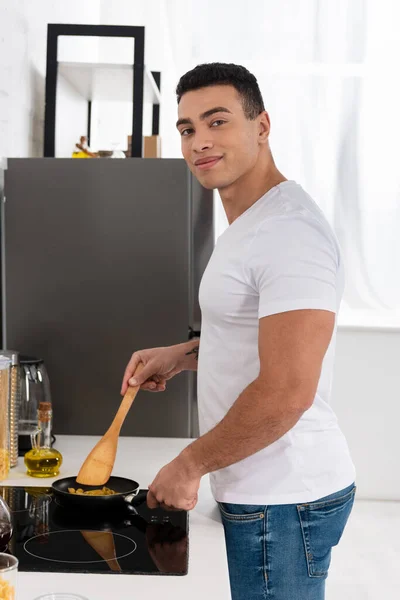 Homem sorrindo, olhando para a câmera e cozinhar com frigideira e espátula perto do fogão da cozinha — Fotografia de Stock