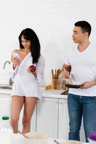 Enfoque selectivo del hombre con sartén y espátula mirando a la chica afroamericana con manzana y teléfono inteligente en la cocina - foto de stock