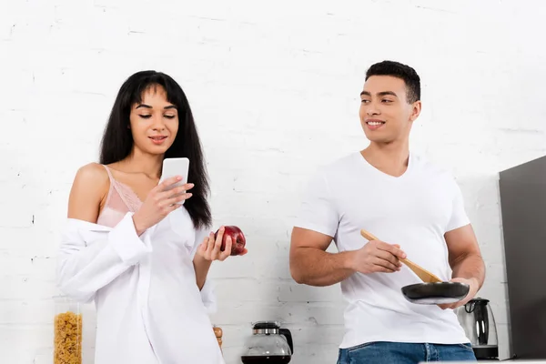 Low angle view of man with frying pan and spatula smiling and looking at african american girl with apple and smartphone — Stock Photo
