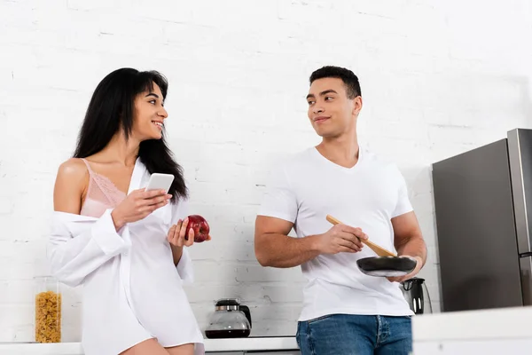 Vista de ángulo bajo de pareja interracial con sartén, espátula, smartphone y manzana mirándose y sonriendo en la cocina - foto de stock