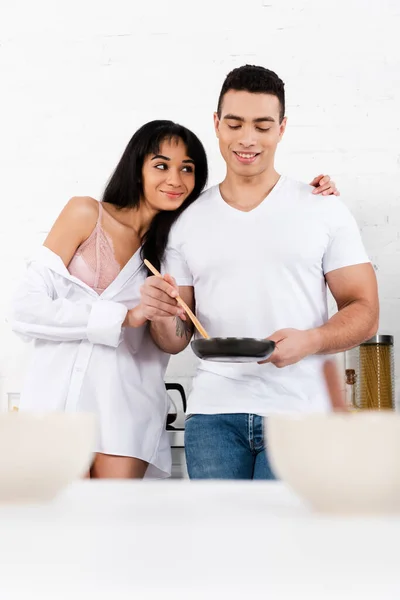 Селективный фокус африканской американской девушки, улыбающейся и обнимающей мужчину сковородкой и лопаткой на кухне — стоковое фото