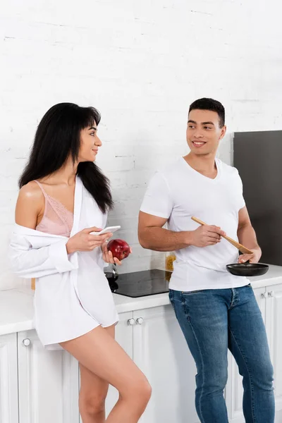 Міжрасові пари зі сковородою, шпателем, смартфоном і яблуком дивляться один на одного і посміхаються на кухні — стокове фото