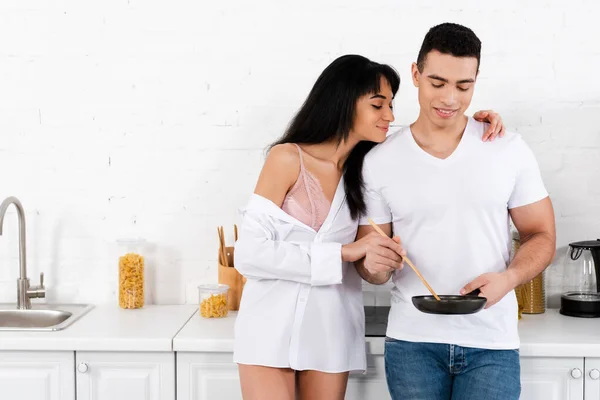 Afroamericano ragazza sorridente e abbracciando l'uomo con padella e spatola in cucina — Foto stock