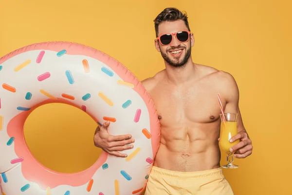 Happy muscular man in sunglasses holding swim ring and cocktail glass with fresh orange juice on yellow background — Stock Photo