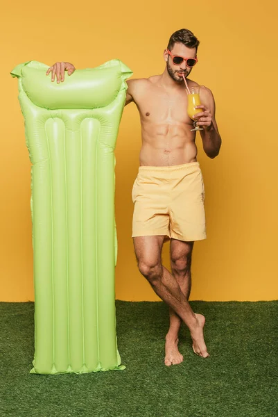 Vista completa de la longitud del hombre sin camisa que bebe el jugo de naranja y que sostiene el colchón inflable mientras que está parado en la hierba verde sobre fondo amarillo - foto de stock