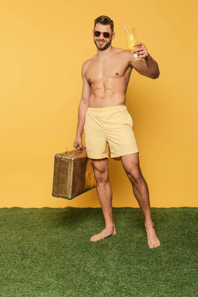 Full length view of happy shirtless man holding cocktail glass with orange juice and vintage suitcase while standing on green grass on yellow background — Stock Photo