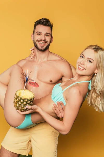 Felice uomo senza maglietta aggrappato mani attraente ragazza bionda con cocktail in ananas su sfondo giallo — Foto stock