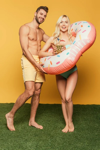 Happy blonde girl in swim ring holding pineapple with cocktail near shirtless muscular man on yellow background — Stock Photo