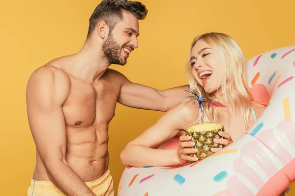 Усміхнений м'язистий чоловік торкається збудженої дівчини в плаванні кільце тримає ананас з коктейлем ізольовано на жовтому — стокове фото