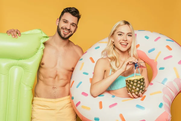 Schöne Shirtess Mann hält aufblasbare Matratze in der Nähe attraktive blonde Mädchen mit Schwimmring und Ananas mit Cocktail isoliert auf gelb — Stockfoto