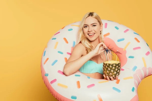 Fröhliches blondes Mädchen im Schwimmring, das die Hälfte der Ananas mit frischem Cocktail auf gelbem Hintergrund hält — Stockfoto