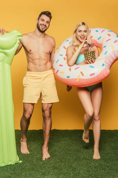 Handsome muscular man with inflatable mattress and smiling blonde girl with swim ring and pineapple with cocktail on yellow background — Stock Photo