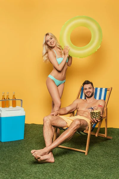 Belle fille blonde avec anneau gonflable et bel homme dans la chaise longue près des bouteilles de bière sur un réfrigérateur portable sur fond jaune — Photo de stock