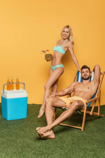 Attraktive blonde Mädchen mit Cocktail in Ananas und schöner Mann im Liegestuhl in der Nähe von Bierflaschen auf tragbarem Kühlschrank auf gelbem Hintergrund — Stockfoto