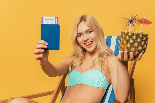 Happy blonde girl in swimsuit holding documents and half of fresh pineapple while sitting in deck chair on yellow background — Stock Photo