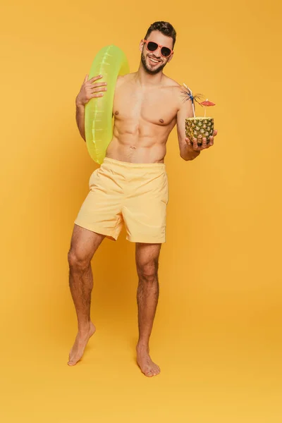 Full length view of happy muscular man in sunglasses holding swim ring and half of pineapple with fresh cocktail on yellow background — Stock Photo