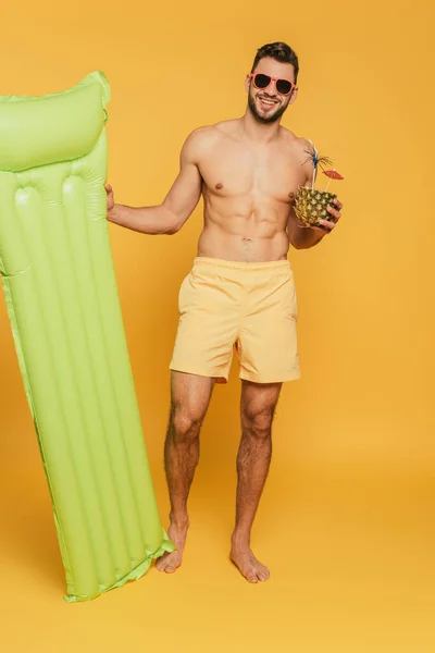 Volle Länge Ansicht des fröhlichen muskulösen Mannes hält aufblasbare Matratze und die Hälfte der Ananas mit frischem Cocktail auf gelbem Hintergrund — Stockfoto