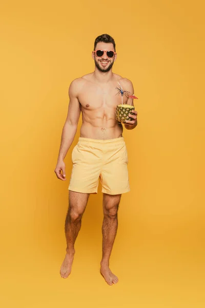 Полный вид на улыбающегося мужчину без рубашки в солнечных очках, держащего половину ананаса со свежим коктейлем на желтом фоне — стоковое фото