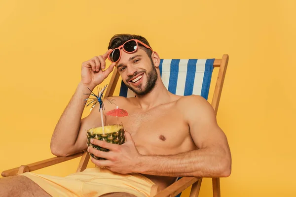 Счастливый мужчина без рубашки дотрагивается до солнцезащитных очков и смотрит в камеру, держа половину ананаса со свежим коктейлем, изолированным на желтом — стоковое фото