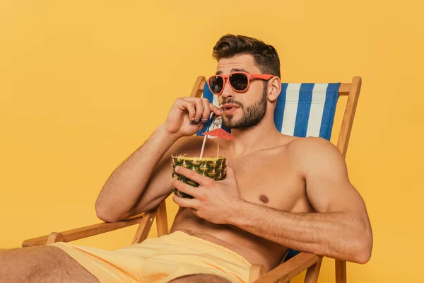 Handsome shirtless man in sunglasses drinking fresh cocktail from half of pineapple while relaxing in deck chair isolated on yellow — Stock Photo
